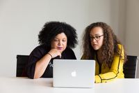 two brown people with curly hair sitting in front of a computer, as the person on the right types