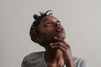 a black person with locs wearing a leopard-spotted turban with their chin resting on their hand and staring upward thoughtfully
