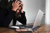 woman with brown hair wearing a black blazer sitting at a computer with her head in her hands, looking confused.