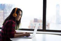 person wearing a red plaid shirt typing at computer while using orange headphones