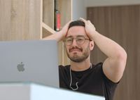 A man wearing glasses staring at his laptop with his head in his hands looking visibly distraught.